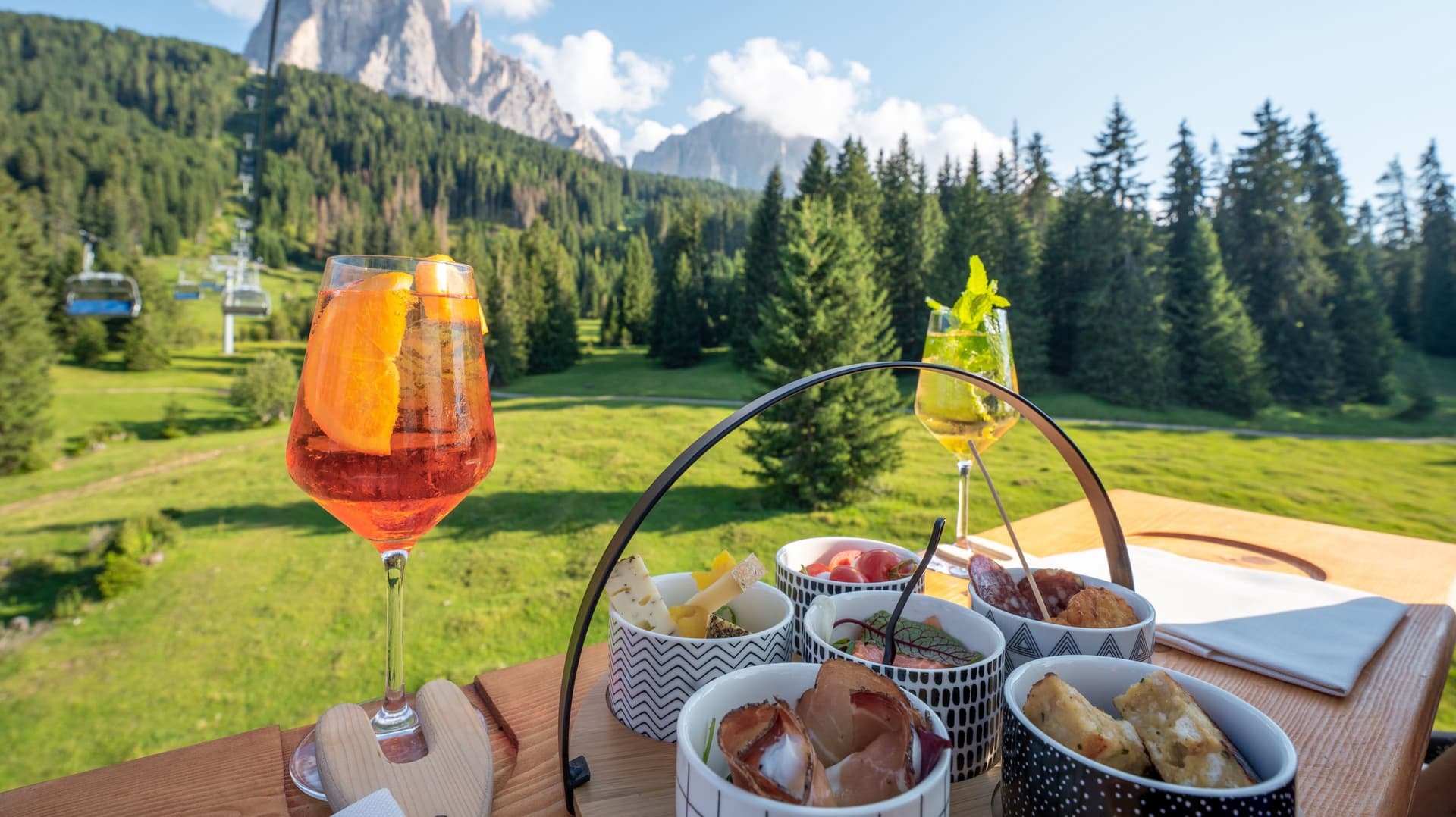 Alzata di antipasti e due calici sul tavolo, dietro la seggiovia che sale tra gli abeti verso il Sassolungo.