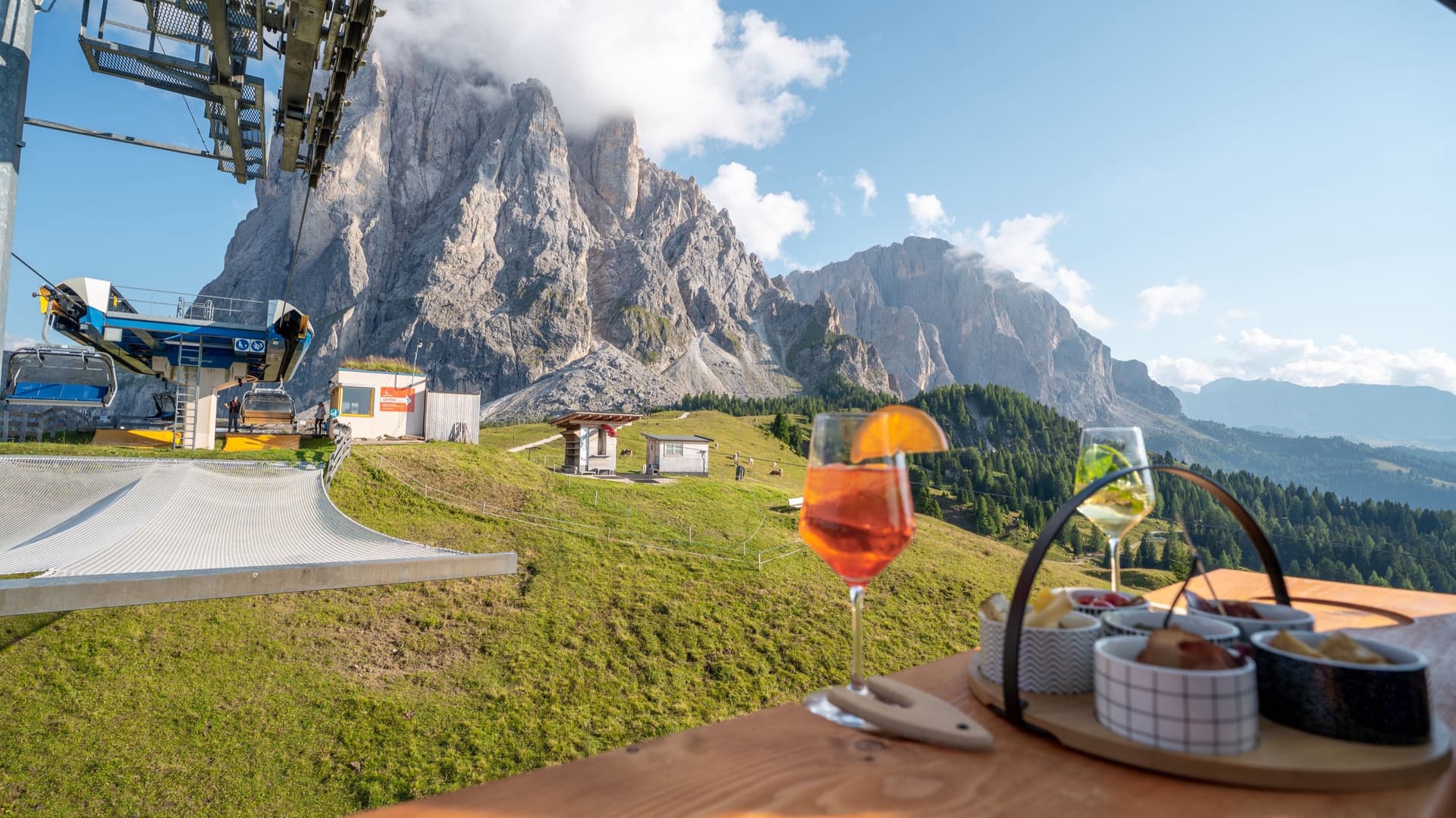 La stazione della seggiovia Mont de Sëura ai piedi del Sassolungo, con due calici e i cestini di antipasti sul tavolo di legno in primo piano.