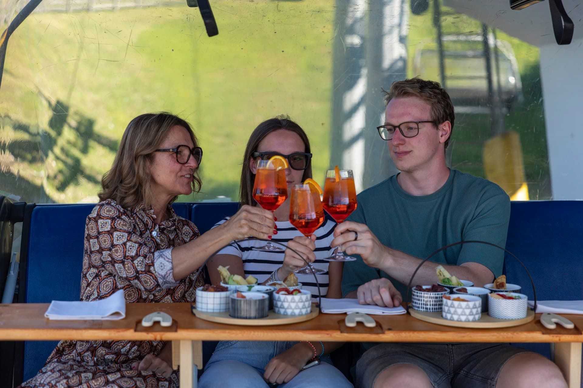 Tre ospiti brindano con lo spritz seduti sulla seggiovia, vassoi di antipasti davanti a loro.